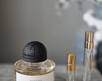 May include: A clear glass perfume bottle with a black textured cap and a white label reading BYREDO BAL D'AFRIQUE ABSOLU DE PARFUM, accompanied by two small, slender, clear glass spray vials with gold-colored caps positioned to the right side.