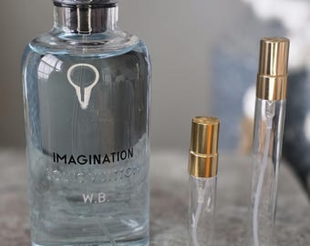 May include: A large clear glass perfume bottle with a black cap, labeled IMAGINATION LOUIS VUITTON W.B., alongside two smaller clear glass spray vials with gold caps. The large bottle contains a light blue liquid and features a white icon.