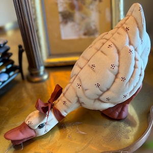 May include: A fabric goose figurine featuring a cream-colored body with a quilted texture and small brown floral accents. The goose has a rust-colored beak and feet, a dark brown ribbon tied around its neck, and painted black eyes with eyelashes.