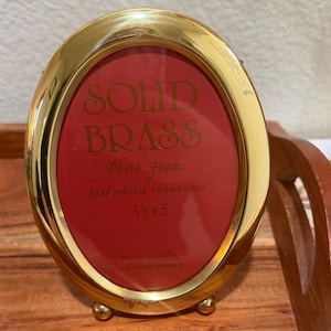 May include: An oval solid brass photo frame with a polished gold finish. The red insert features the text: SOLID BRASS Photo Frame hand polished lacquer coated 3 1/2 x 5 by Intercraft THE FRAME PEOPLE. The frame stands on two small metal feet.