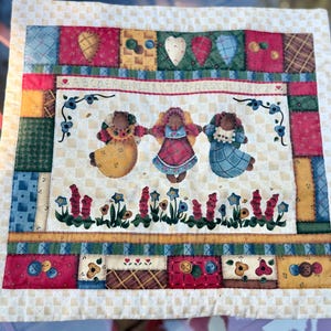May include: A square fabric wall hanging featuring a patchwork border with red, green, blue, and yellow squares. The center panel displays three bear figures in dresses, an alphabet sequence, and floral motifs on a cream checkered background.