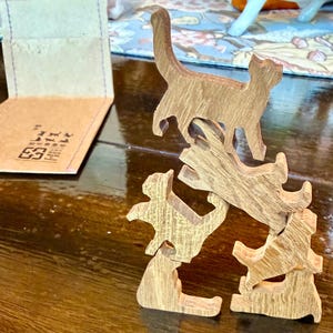 May include: A set of five interlocking wooden cat figurines stacked in a vertical tower formation. Each piece is carved from light brown wood with a visible grain pattern, depicting cats in various standing, sitting, and stretching poses.