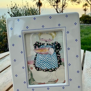 May include: A white ceramic picture frame with a blue border and small blue cross patterns. The frame holds an illustration of a person in a dark floral dress and blue checkered apron holding a bowl, surrounded by white geese in a grassy area.