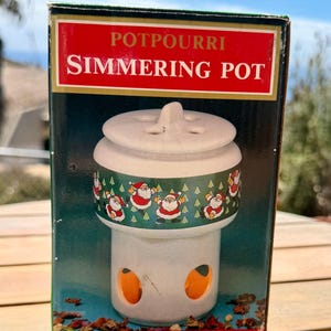 May include: A rectangular product box for a potpourri simmering pot. The box features a white ceramic pot with a lid, decorated with a green band showing Santa Claus figures and pine trees. The text reads POTPOURRI SIMMERING POT in white letters.