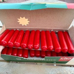 May include: A cardboard box containing forty-eight red cylindrical candles arranged in two rows. The green box features the text "ADVENTKERZEN" and the brand name "eika" in white and red. The interior of the lid has a star-shaped cutout.