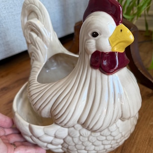 May include: A cream-colored ceramic chicken-shaped planter or container with a glossy finish. The figure features a bright yellow beak, a deep red comb, and dark red wattles. The body has molded feather textures and a hollow opening at the top.