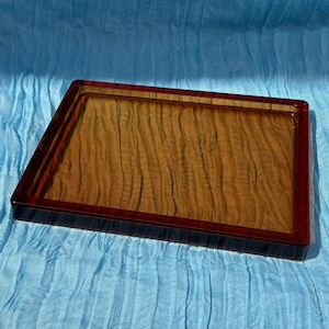 Puede incluir: Una bandeja rectangular con un borde de color ámbar intenso y un panel central con un patrón de vetas de madera marrón claro. La bandeja tiene un acabado liso y brillante y un borde elevado, presentando un diseño funcional y limpio.