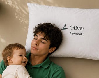 Könnte beinhalten: Ein rechteckiges weißes Kissen mit einem dunkelgrünen Blattsymbol, gefolgt von dem in Schwarz gedruckten Text Oliver und 2 years old. Das Kissen hat eine glatte, einfarbig weiße Oberfläche und liegt horizontal auf einem neutralfarbenen Untergrund.