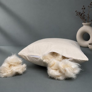May include: A square, off-white cotton pillow cover with a visible zipper opening. A portion of the soft, cream-colored natural fiber stuffing is pulled out through the open zipper, and a separate clump of the same fibrous material sits nearby.