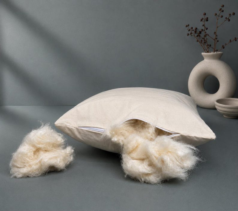 May include: A square, off-white cotton pillow cover with a visible zipper opening. A portion of the soft, cream-colored natural fiber stuffing is pulled out through the open zipper, and a separate clump of the same fibrous material sits nearby.