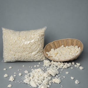 Op de afbeelding: Een doorzichtige plastic zak en een rieten mand gevuld met witte, versnipperde schuim. Enkele schuimstukken liggen verspreid op een grijze ondergrond. Het schuim lijkt een kussen- of kussenvulling te zijn.