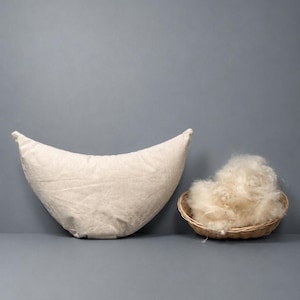 May include: A cream-colored, crescent-shaped pillow and a small woven basket filled with fluffy, light-colored material. The pillow is made of a textured fabric. The basket is round and made of woven material.