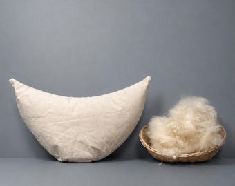 Könnte beinhalten: Ein cremefarbenes, halbmondförmiges Kissen und ein kleiner Weidenkorb, gefüllt mit flauschigem, hellfarbenem Material. Das Kissen besteht aus einem strukturierten Stoff. Der Korb ist rund und aus geflochtenem Material.