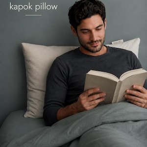 May include: A rectangular off-white kapok pillow with a small fabric tag on the side. The text "kapok pillow" is printed in white above the pillow, accompanied by a simple line-drawn leaf icon and a horizontal line separator.