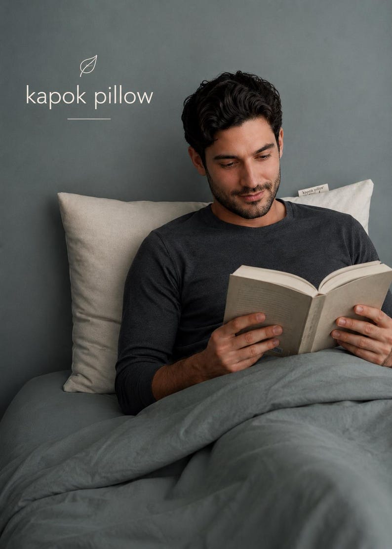 May include: A rectangular off-white kapok pillow with a small fabric tag on the side. The text "kapok pillow" is printed in white above the pillow, accompanied by a simple line-drawn leaf icon and a horizontal line separator.
