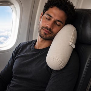 May include: A cylindrical travel pillow in a light beige, off-white fabric with a visible side zipper and a small fabric tag. The pillow is positioned against the side of a dark gray airplane seat, providing support for the head and neck area.