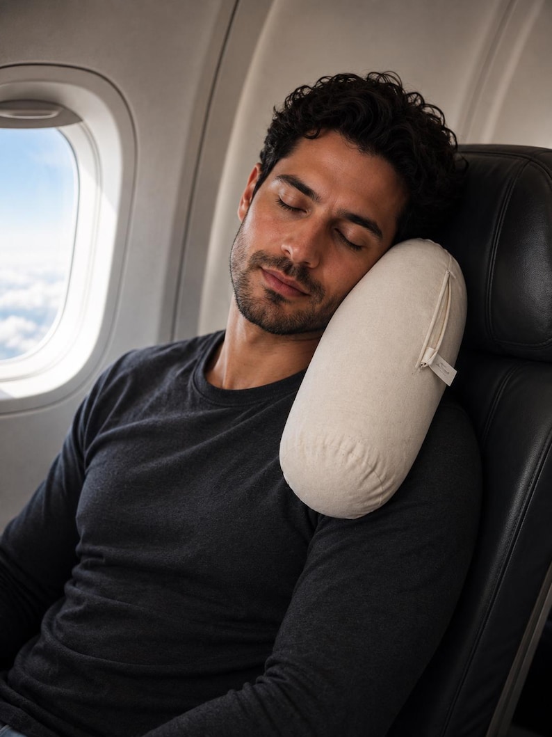 May include: A cylindrical travel pillow in a light beige, off-white fabric with a visible side zipper and a small fabric tag. The pillow is positioned against the side of a dark gray airplane seat, providing support for the head and neck area.