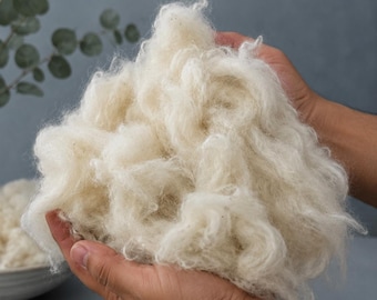 Könnte beinhalten: Ein großer, flauschiger Haufen aus roher, cremeweißer Wollfaser mit einer weichen, gekräuselten Textur. Das lose, ungesponnene Material wird in den Händen gehalten und zeigt das natürliche, luftige Volumen und die faserige Beschaffenheit.