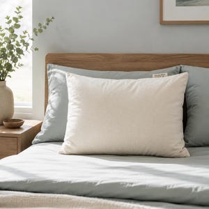 May include: A rectangular off-white pillow with a smooth, solid-colored fabric cover. A small, rectangular fabric tag is attached to the top right corner of the pillow, featuring the printed text "kapok pillow" in a simple, dark font.