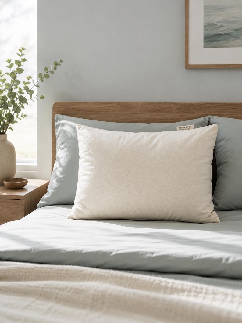May include: A rectangular off-white pillow with a smooth, solid-colored fabric cover. A small, rectangular fabric tag is attached to the top right corner of the pillow, featuring the printed text "kapok pillow" in a simple, dark font.
