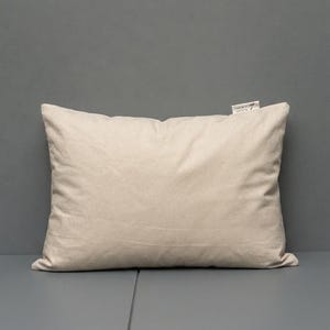 May include: A rectangular throw pillow in a solid, light beige color. The pillow is made of a plain, woven fabric and features a small, white fabric tag attached to the top right corner with black printed text that reads 'JUNE BY 7' and 'MADE IN TR'.