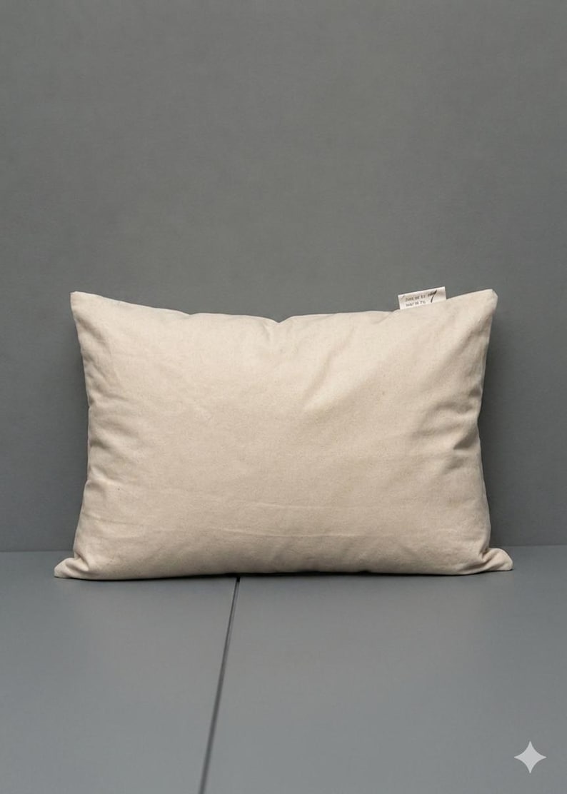 May include: A rectangular throw pillow in a solid, light beige color. The pillow is made of a plain, woven fabric and features a small, white fabric tag attached to the top right corner with black printed text that reads 'JUNE BY 7' and 'MADE IN TR'.