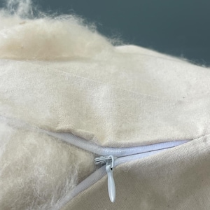 May include: A beige fabric pillow cover featuring a white zipper closure. The pillow is partially open, revealing a soft, fluffy, off-white fibrous filling material that extends outward from the opening of the textile casing.