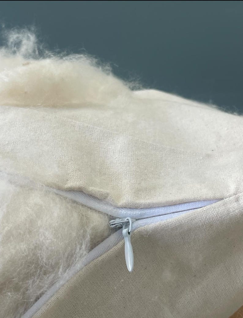 May include: A beige fabric pillow cover featuring a white zipper closure. The pillow is partially open, revealing a soft, fluffy, off-white fibrous filling material that extends outward from the opening of the textile casing.