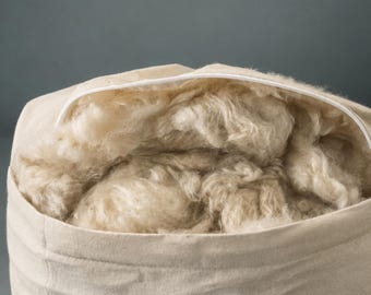 Könnte beinhalten: Ein beiger Stoffbeutel gefüllt mit flauschiger, hellfarbener Wolle. Der Beutel hat einen weißen Reißverschluss und ist vor einem gedämpften grauen Hintergrund platziert. Die Wolle wirkt weich und voluminös.