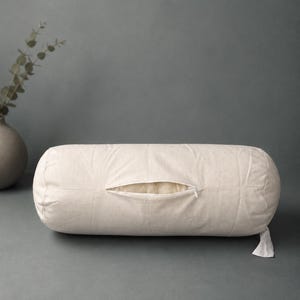 May include: A cylindrical bolster pillow in an off-white, textured fabric. The pillow features a central horizontal zipper opening that is partially unzipped, revealing a soft, light-colored interior lining. A small fabric tag hangs from the side.