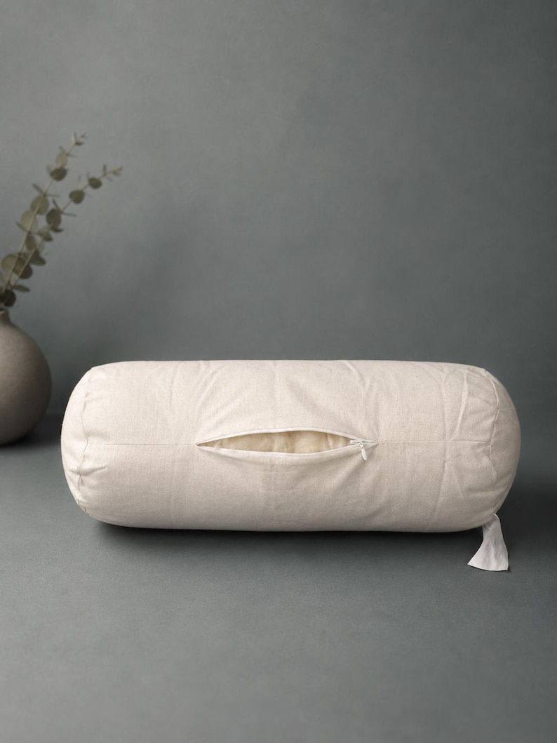 May include: A cylindrical bolster pillow in an off-white, textured fabric. The pillow features a central horizontal zipper opening that is partially unzipped, revealing a soft, light-colored interior lining. A small fabric tag hangs from the side.