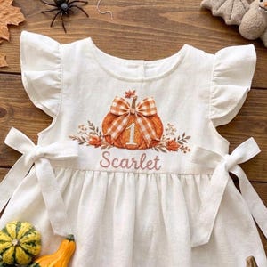 May include: A white dress with flutter sleeves and side ties, featuring an embroidered orange pumpkin with a plaid bow, the number 1, and the name Scarlet in cursive script. The design includes autumn leaves and vines surrounding the pumpkin.