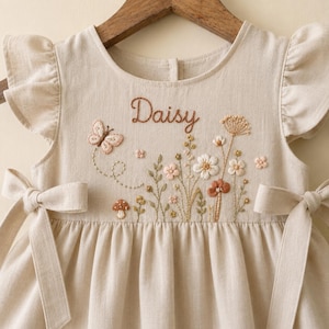 May include: A beige dress with ruffled sleeves and side waist ties. The bodice features brown embroidered text reading Daisy, accompanied by detailed embroidery of a butterfly, various wildflowers, and a small mushroom in soft earth tones.