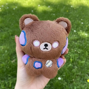 May include: A small brown plush bear toy with soft fur, featuring embroidered light purple patches on the ears, shoulders, and chest. The bear has white circular eyes, a black nose, and a small silver-colored stitched detail on its chest.
