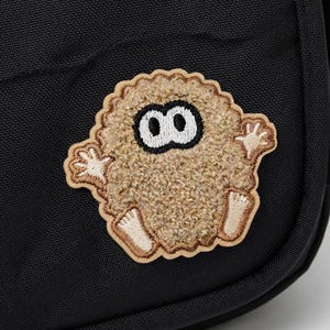 May include: A black bag featuring a textured, beige, fuzzy creature patch with large white eyes and black pupils. The creature has small arms and feet. A black fabric tag on the side displays the white text NINTENDO TOKYO JAPAN in capital letters.