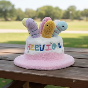 May include: A pink and white textured bucket hat featuring embroidered colorful letters that spell out HEEVIOU and small star accents. The top of the hat is adorned with several plush, worm-like figures in shades of yellow, blue, purple, and pink.