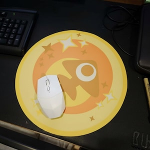 May include: A white wireless computer mouse rests on a round yellow mousepad with a rocket ship design. The mousepad has a star and circle design in shades of yellow and orange. A black keyboard is visible in the background.
