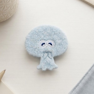 May include: A light blue felted patch shaped like a stylized jellyfish or mushroom character. The patch features a rounded top and a smaller base with two embroidered white oval eyes containing dark blue curved pupils, creating a simple face.