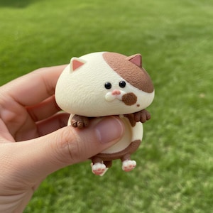 May include: A small, adorable cat figurine with a cream-colored body and brown markings. The cat has small pink ears, black eyes, and a brown spot on its face. Its paws are also brown and pink. The figurine is held against a green background.