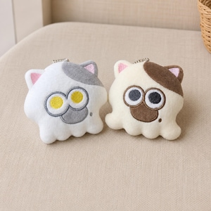 May include: Two plush cat-shaped keychains with metal ball chains. One is white with a gray patch and yellow eyes, the other is cream with a brown patch and black eyes. Both feature pink inner ears and a small embroidered dot on the cheek.