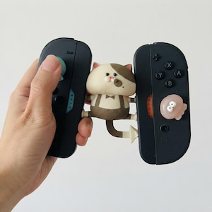 May include: A pair of dark gray gaming controllers connected by a central figurine of a cream and brown cat wearing brown suspenders and a bow tie. The left controller features a light blue joystick cover, while the right has a pink octopus cover.