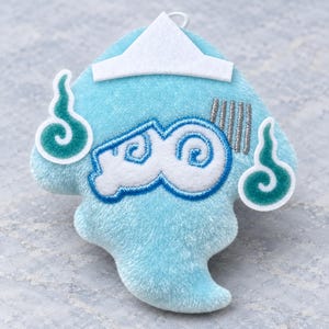 May include: A light blue plush character ornament featuring a white paper-style hat, two teal swirl accents on the sides, and a central white embroidered design with blue outlines. The character has a soft, fuzzy texture and a small white loop.