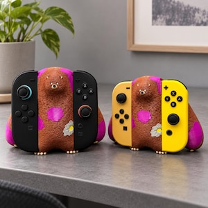 May include: Two brown bear-shaped game controller stands with textured fur, pink patches, and a white flower detail. One stand holds black controllers with blue and orange accents, while the other holds yellow controllers with black buttons.