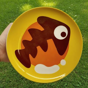 May include: A round yellow plate featuring a central orange circle and a dark brown graphic of a fish skeleton. The design includes a large white eye on the fish head and a white splash shape at the bottom, creating a bold, high-contrast visual.
