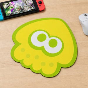Puede incluir: Una alfombrilla de ratón de color amarillo y verde lima con forma de personaje de calamar con dos grandes ojos blancos y pupilas verdes. El diseño incluye una serie de pequeños puntos circulares a lo largo del borde inferior, imitando una base de tentáculo.