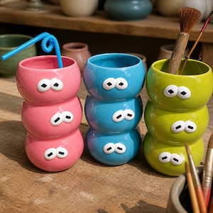 May include: Three stacked ceramic cups in pink, blue, and lime green, each featuring three pairs of white eyes with black pupils. The pink cup contains a blue plastic straw, while the lime green cup holds a wooden-handled brush.