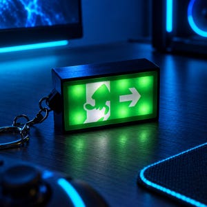May include: A rectangular black keychain featuring a glowing green emergency exit sign. The sign displays a white running figure icon and a white arrow pointing to the right. The keychain includes a silver metal ring attached by a short chain.