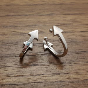 May include: A pair of silver-toned metal ear cuffs shaped like arrows. Each cuff features a pointed arrow head at one end and a small spherical ball at the other, with a secondary arrow-shaped detail positioned along the curved band of the jewelry.