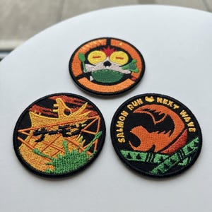 May include: Three circular embroidered patches with black borders. The top patch features a yellow-eyed character. The bottom left patch shows a yellow structure with black Japanese text. The bottom right patch displays an orange icon and the text SALMON RUN NEXT WAVE.