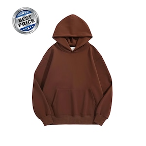 Puede incluir: Una sudadera con capucha de color marrón sólido que presenta mangas largas, un bolsillo tipo canguro frontal y puños y dobladillo acanalados. Una insignia plateada circular muestra el texto BEST PRICE con estrellas y GUARANTEED.